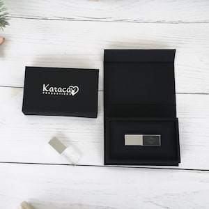以下が含まれることがあります： 黒い箱の中に銀色のUSBドライブが入っています。箱の上には「Karaca Productions」という文字が書かれています。箱は白い木製の表面の上に置かれています。