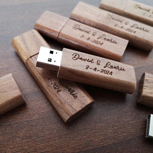 Op de afbeelding: Houten USB-sticks met de tekst "David & Laurie 2-4-2024" gegraveerd. De sticks zijn lichtbruin van kleur en hebben een natuurlijke houtnerf.