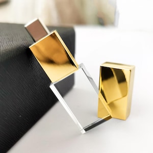May include: A rectangular clear glass perfume bottle with a reflective gold metal cap and a gold metal collar. The bottle features a sleek, minimalist design with sharp, clean edges and a transparent body that allows for a clear view of the interior.