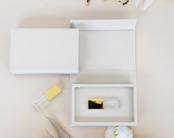 Könnte beinhalten: Eine weiße Leinenbox mit einem goldenen USB-Stick im Inneren. Die Box ist geöffnet und der USB-Stick ist sichtbar. Der USB-Stick ist klar mit einer goldenen Oberseite.
