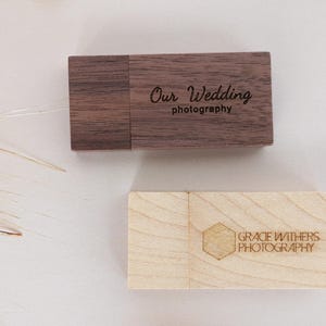 Puede incluir: Dos unidades USB de madera. La primera unidad es de madera de nogal con el texto "Our Wedding Photography" grabado en ella. La segunda unidad es de madera de arce con el texto "Grace Withers Photography" grabado en ella.