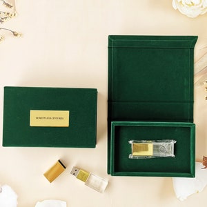 Puede incluir: Dos cajas de terciopelo verde con letras doradas. La caja más pequeña contiene una unidad USB dorada y transparente. La caja más grande tiene el texto "Moments for Centuries" en la parte delantera.