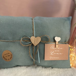 Könnte beinhalten: Ein teal-farbenes Kissen, das mit Bindfaden und einem hellblauen Band umwickelt ist, verziert mit einem Holzherz und einem Etikett mit der Aufschrift "Made with Love". Ein kleiner, runder Aufkleber mit den Worten "handmade with love" ist ebenfalls sichtbar.