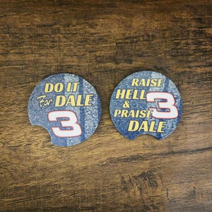 May include: Two blue car coasters with white and yellow text. One says "DO IT For DALE 3" and the other says "RAISE HELL & PRAISE DALE 3". The coasters have a textured, speckled design.