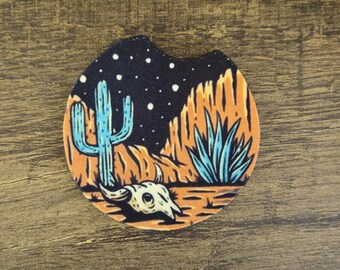 Puede incluir: Posavasos redondo para coche con diseño de paisaje desértico. El posavasos representa un cactus azul, formaciones rocosas naranjas y una calavera bajo un cielo estrellado. El posavasos está diseñado para caber en el portavasos de un coche.