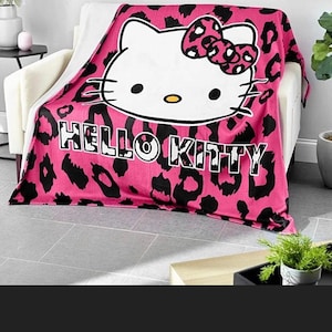 May include: Pink and black Hello Kitty blanket. The blanket features a large Hello Kitty face with a bow and the words "HELLO KITTY" in black block letters. The background has a pink leopard print.