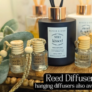 May include: A collection of reed diffusers in glass bottles with wooden caps and gold string. The bottles are labeled with different scents, including "Pacific Peaks", "Sun Kissed", "Desert Blooms", and "Sweet & Spicy". The text "Reed Diffusers" and "hanging diffusers also available" is displayed below the bottles.