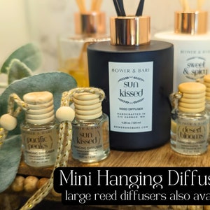 May include: Four mini hanging reed diffusers with wooden caps and gold cords. The scents are "Pacific Peaks", "Sun Kissed", "Desert Blooms", and "Sweet & Spicy". A larger black reed diffuser bottle with the scent "Sun Kissed" is also shown. The text "Mini Hanging Diffusers" and "Large Reed Diffusers Also Available" is displayed below the image.