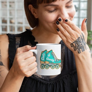 Può includere: Una tazza in ceramica bianca con un'immagine di pattini a rotelle color turchese e il testo "Cute & Gunky".