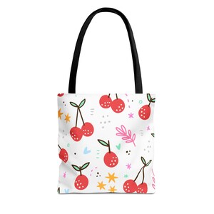 May include: White tote bag with a repeating pattern of red cherries, green leaves, and colorful stars. The bag has black handles and is designed for carrying items. The cherry design is whimsical and playful.