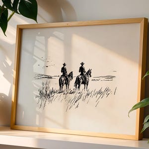 K&ouml;nnte beinhalten: Ein gerahmter Schwarz-Wei&szlig;-Druck, der zwei Cowboys auf Pferden in einem Feld zeigt. Das Kunstwerk befindet sich in einem Holzrahmen und lehnt an einer wei&szlig;en Oberfl&auml;che. Die Cowboys tragen H&uuml;te und reiten auf Pferden.