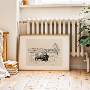 K&ouml;nnte beinhalten: Ein gerahmter Schwarz-Wei&szlig;-Druck eines Bisons auf einem Feld. Das Kunstwerk befindet sich in einem hellen Holzrahmen und steht auf einem hellen Holzboden. Links vom Kunstwerk befinden sich ein Stapel B&uuml;cher und ein Handtuchhalter aus Holz.