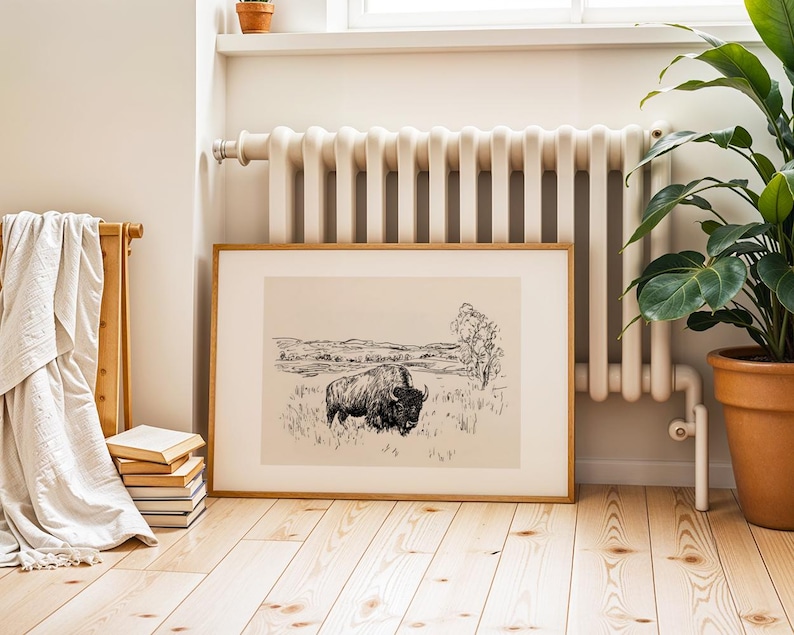 K&ouml;nnte beinhalten: Ein gerahmter Schwarz-Wei&szlig;-Druck eines Bisons auf einem Feld. Das Kunstwerk befindet sich in einem hellen Holzrahmen und steht auf einem hellen Holzboden. Links vom Kunstwerk befinden sich ein Stapel B&uuml;cher und ein Handtuchhalter aus Holz.