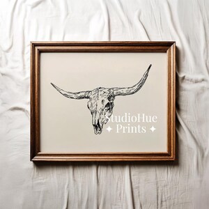K&ouml;nnte beinhalten: Ein gerahmter Druck mit einer schwarz-wei&szlig;en Illustration eines Longhorn-Sch&auml;dels. Das Kunstwerk ist vor einem neutralen Hintergrund platziert und in dunklem Holz gerahmt. Der Text "studioHue Prints" ist sichtbar.