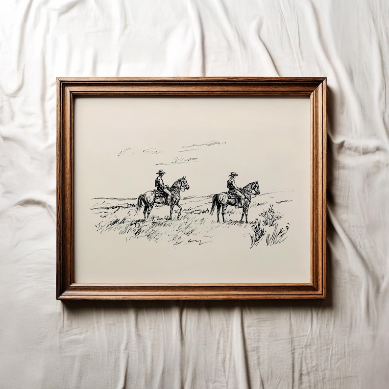 May include: A framed black and white print depicting two cowboys on horseback in a grassy field. The artwork is encased in a dark brown wooden frame, adding a rustic touch to the western-themed illustration.