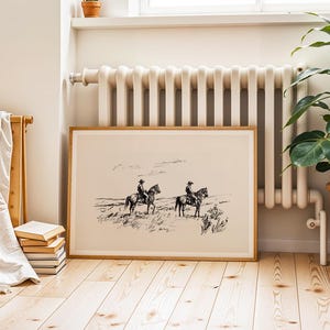 May include: A framed black and white print depicting two cowboys on horseback in a landscape. The artwork is in a light wood frame and rests on a wooden floor. A radiator and a potted plant are in the background.
