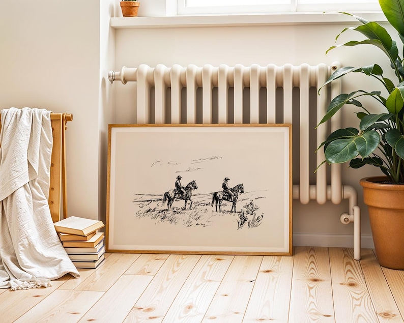 May include: A framed black and white print depicting two cowboys on horseback in a landscape. The artwork is in a light wood frame and rests on a wooden floor. A radiator and a potted plant are in the background.