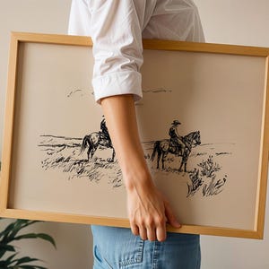 May include: A framed print featuring a black and white sketch of two cowboys on horseback in a grassy field. The artwork has a beige background and is encased in a light brown wooden frame. The image evokes a vintage, rustic aesthetic.