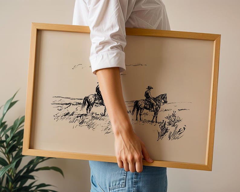 May include: A framed print featuring a black and white sketch of two cowboys on horseback in a grassy field. The artwork has a beige background and is encased in a light brown wooden frame. The image evokes a vintage, rustic aesthetic.