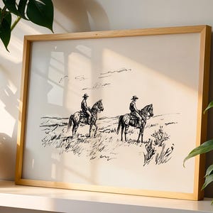 May include: A framed black and white print featuring two cowboys on horseback in a grassy field. The artwork is in a wooden frame and has a minimalist, sketched style. The print evokes a Western theme.
