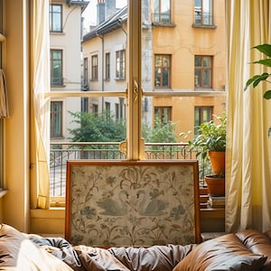 May include: A framed artwork featuring two swans amidst floral patterns, displayed in front of a window overlooking a European cityscape. The room has tan curtains, a brown leather couch, and potted plants.