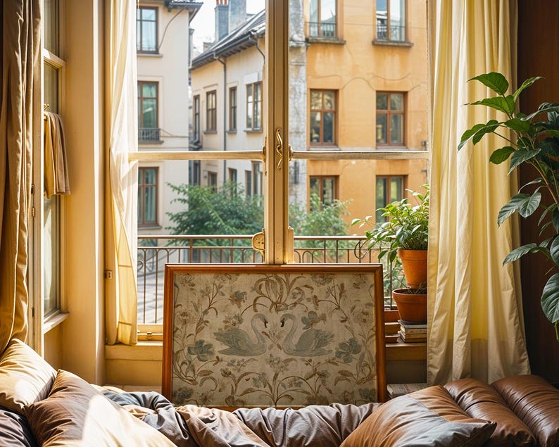 May include: A framed artwork featuring two swans amidst floral patterns, displayed in front of a window overlooking a European cityscape. The room has tan curtains, a brown leather couch, and potted plants.
