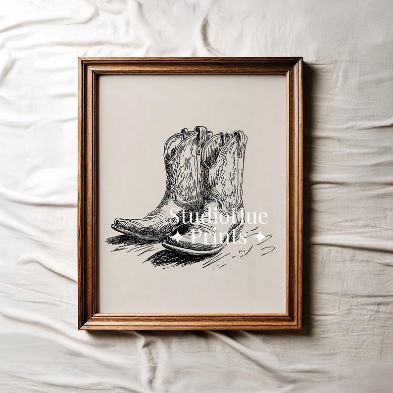 K&ouml;nnte beinhalten: Ein gerahmter Schwarz-Wei&szlig;-Druck mit einer detaillierten Illustration eines Paares von Cowboy-Stiefeln. Die Stiefel sind mit Schattierungen und Texturen versehen, vor einem neutralen Hintergrund. Der Rahmen hat einen warmen, nat&uuml;rlichen Holzton.