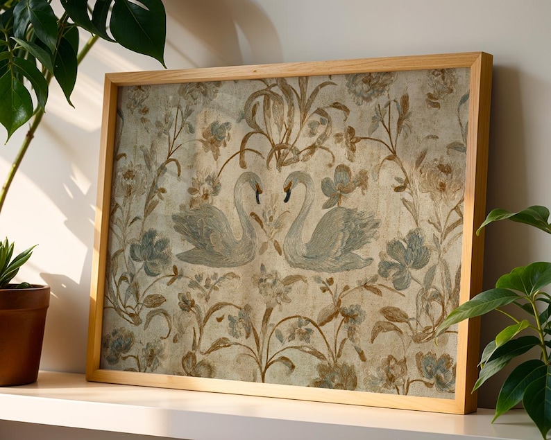 May include: A framed artwork featuring a vintage-style design. The artwork showcases two swans facing each other, surrounded by floral and botanical elements in muted blue, brown, and beige tones. The frame is made of light-colored wood.