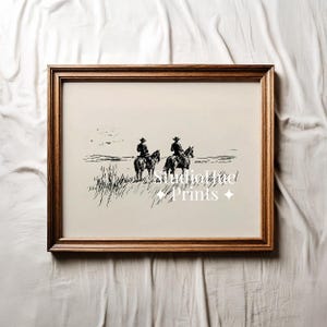 K&ouml;nnte beinhalten: Ein gerahmter Schwarz-Wei&szlig;-Druck, der zwei Cowboys auf Pferden in einem grasbewachsenen Feld zeigt. Das Kunstwerk hat eine Vintage-&Auml;sthetik mit dem Text "StudioHut Prints" in der Mitte. Der Rahmen hat eine warme braune Farbe.