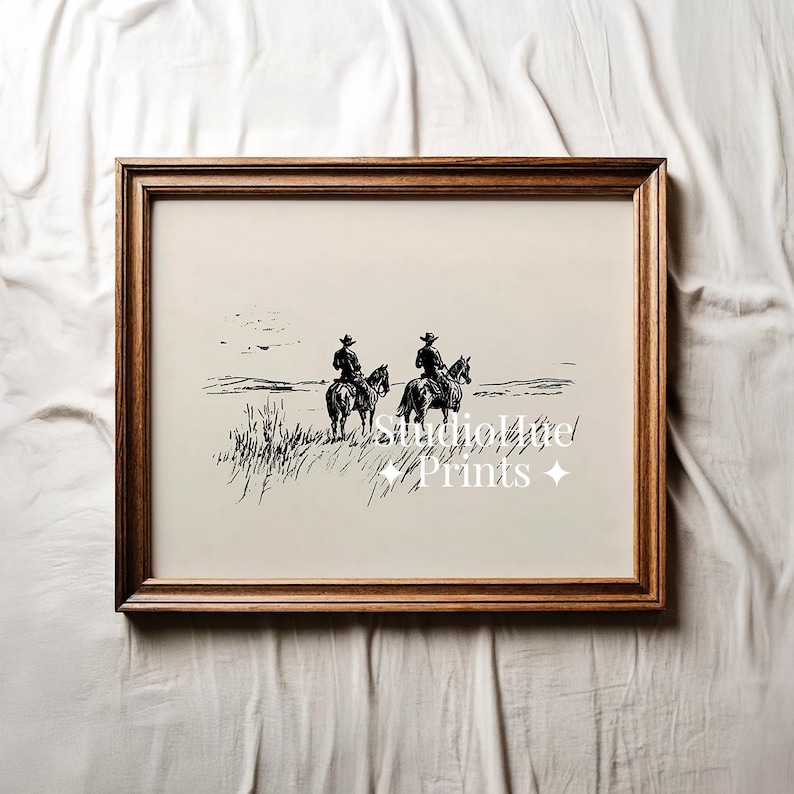 K&ouml;nnte beinhalten: Ein gerahmter Schwarz-Wei&szlig;-Druck, der zwei Cowboys auf Pferden in einem grasbewachsenen Feld zeigt. Das Kunstwerk hat eine Vintage-&Auml;sthetik mit dem Text "StudioHut Prints" in der Mitte. Der Rahmen hat eine warme braune Farbe.