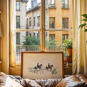May include: A framed black and white print of two cowboys on horseback in a landscape scene. The artwork is displayed on a bed with brown leather cushions, near a window with yellow curtains and a view of a European-style building.