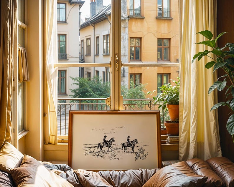 May include: A framed black and white print of two cowboys on horseback in a landscape scene. The artwork is displayed on a bed with brown leather cushions, near a window with yellow curtains and a view of a European-style building.