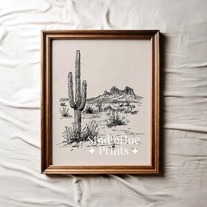 K&ouml;nnte beinhalten: Gerahmter Schwarz-Wei&szlig;-Kunstdruck einer W&uuml;stenlandschaft mit einem Saguaro-Kaktus, Bergen und W&uuml;stenpflanzen. Das Kunstwerk befindet sich in einem Holzrahmen und enth&auml;lt den Text "Studio Hue Prints".