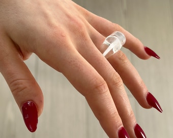 May include: A clear plastic ring with a unique design. The ring is worn on a finger with red nail polish.