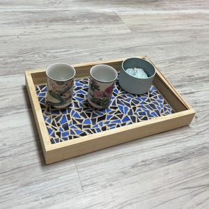 May include: A wooden serving tray with a mosaic tile design in shades of blue and black. The tray holds two decorative teacups with floral patterns and a small blue container. The tray is made of light-colored wood.