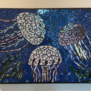 Puede incluir: Una obra de arte en mosaico que presenta tres medusas sobre un fondo azul oscuro. Las medusas están hechas con teselas blancas, moradas e iridiscentes, con tentáculos colgantes. El mosaico está enmarcado en negro, creando un contraste llamativo.