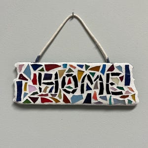 May include: A rectangular mosaic sign with the word "HOME" in black letters. The sign is made of colorful glass pieces in various shapes and sizes, set against a white background. A white rope is attached for hanging.