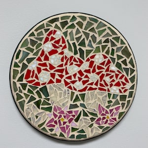 May include: Round mosaic art piece featuring a mushroom design. The mushrooms are red with white spots, set against a green background. The mosaic includes pink flowers at the bottom and is framed with a black border.