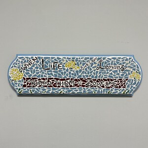 May include: A mosaic-style sign with a light blue border and background. The sign reads "When life gives you lemons, make someone" in a mix of colors. Yellow lemons are depicted on the left and right sides of the sign.