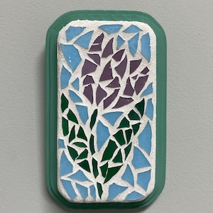 Könnte beinhalten: Ein rechteckiges Mosaikkunstwerk mit floralem Design. Das Mosaik besteht aus blauen, violetten und grünen Glasstücken auf weißem Hintergrund, umrahmt von einem türkisgrünen Rand. Das Kunstwerk ist an einer hellgrauen Wand befestigt.