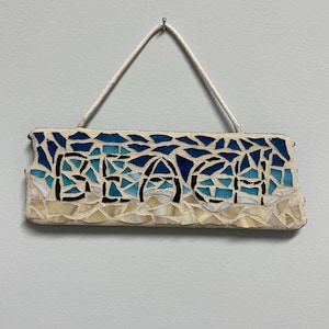May include: A rectangular mosaic sign with the word "BEACH" in black letters. The top of the sign features blue mosaic tiles, transitioning from dark to light, resembling the ocean. The bottom has beige tiles, suggesting sand. A white rope is used for hanging.