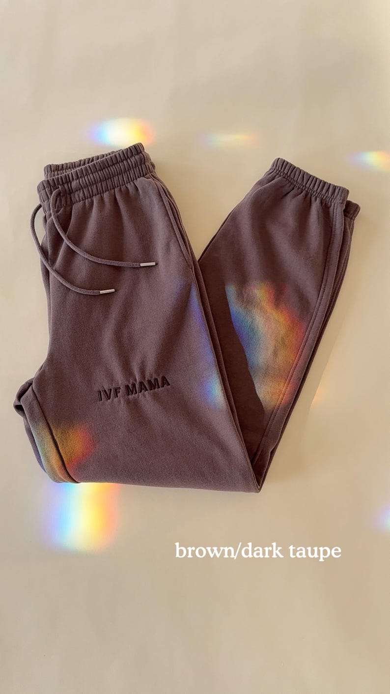 Puede incluir: Un pantal&oacute;n de ch&aacute;ndal marr&oacute;n/taupe oscuro con el texto "IVF MAMA" bordado en la pierna izquierda.