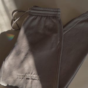 Puede incluir: Pantalones de ch&aacute;ndal de color verde kaki claro con el texto "IVF Mama" bordado en la pierna izquierda.