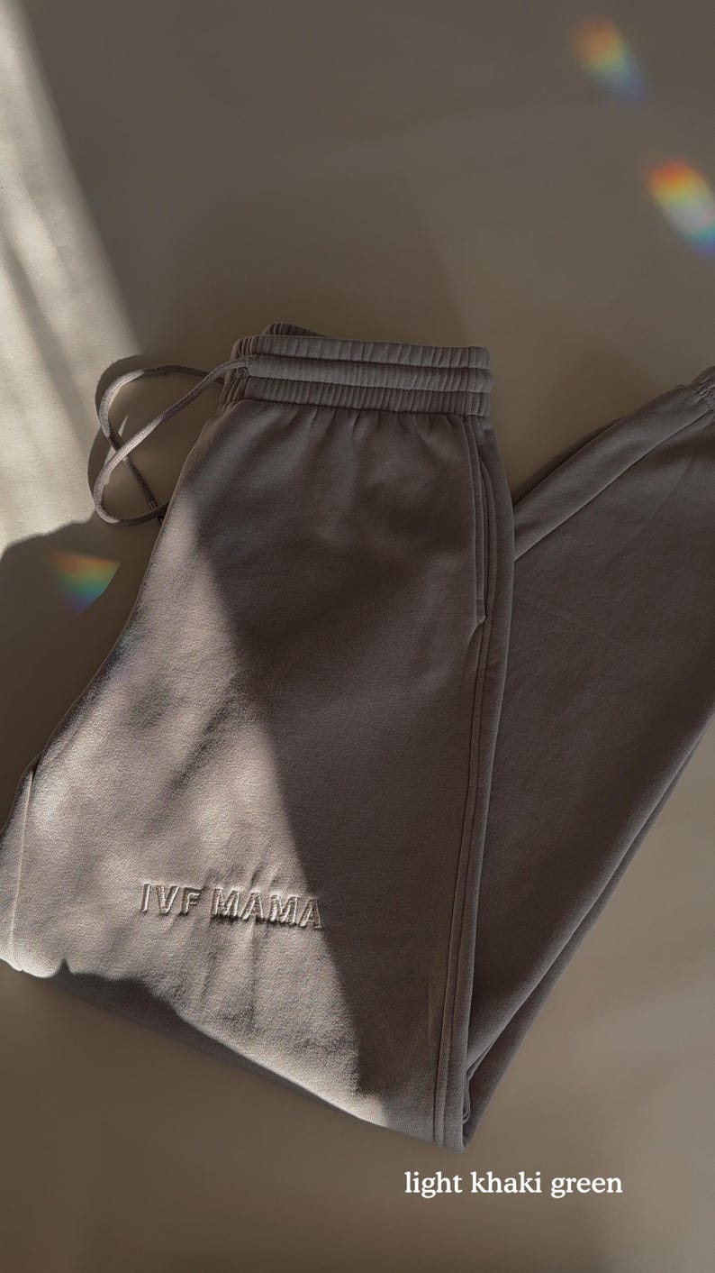 Puede incluir: Pantalones de ch&aacute;ndal de color verde kaki claro con el texto "IVF Mama" bordado en la pierna izquierda.