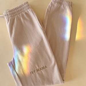 Puede incluir: Un par de pantalones de ch&aacute;ndal de color claro con el texto "IVF MAMA" bordado en la pierna izquierda. Los pantalones de ch&aacute;ndal est&aacute;n colocados sobre una superficie de color crema.