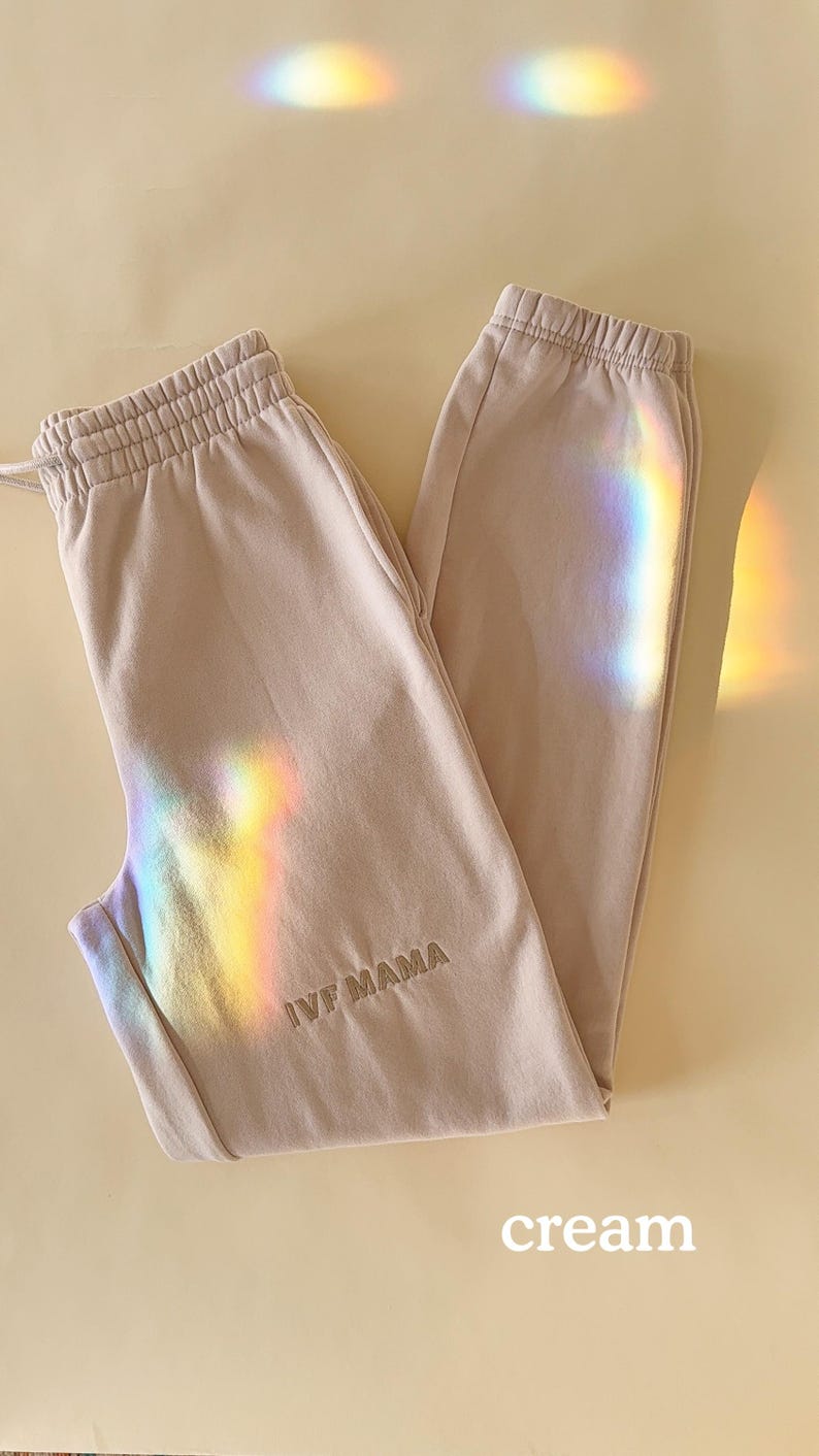 Puede incluir: Un par de pantalones de ch&aacute;ndal de color claro con el texto "IVF MAMA" bordado en la pierna izquierda. Los pantalones de ch&aacute;ndal est&aacute;n colocados sobre una superficie de color crema.