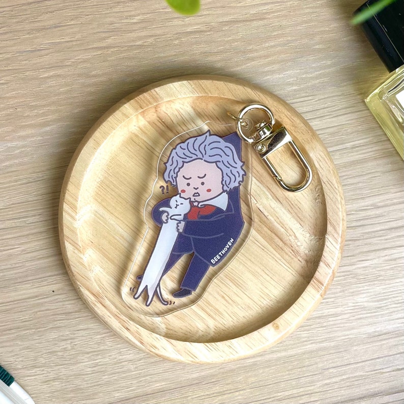 May include: A clear acrylic keychain featuring a cartoon illustration of Ludwig van Beethoven holding a white cat. The cat is looking at Beethoven with a surprised expression. The keychain has a gold metal ring and clasp. The text "BEETHOVEN" is printed on the keychain.
