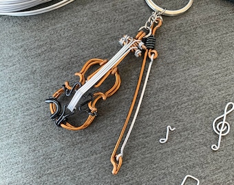 May include: A silver wire violin keychain with a brown wire body and a silver wire bow. The keychain is attached to a silver key ring. There are three silver wire music notes on a gray background.