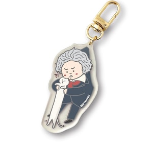 May include: A clear acrylic keychain featuring a cartoon illustration of Ludwig van Beethoven holding a white cat. The cat is looking up at Beethoven, who has a surprised expression on his face. The text "BEETHOVEN" is printed on the keychain.