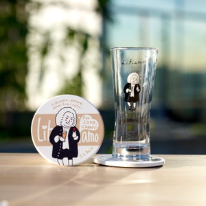 May include: A white ceramic coaster with a cartoon illustration of a man in a black suit and white wig holding a glass of wine. The coaster has the text "Libiamo, amore. amor fra i calisi. Lit jamo J.S. Bach" printed on it. The coaster is next to a clear glass with the text "Libiamo" printed on it.